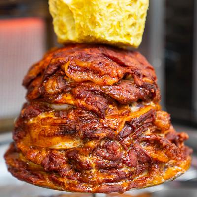 A vertical rotisserie spit cooking al pastor with a piece of pineapple placed on top.