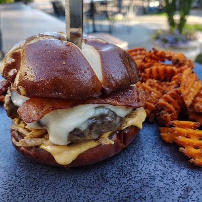 Bacon cheeseburger on a glossy pretzel bun served with waffle fries outdoors.