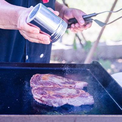 Chef seasoning a steak on a grill.