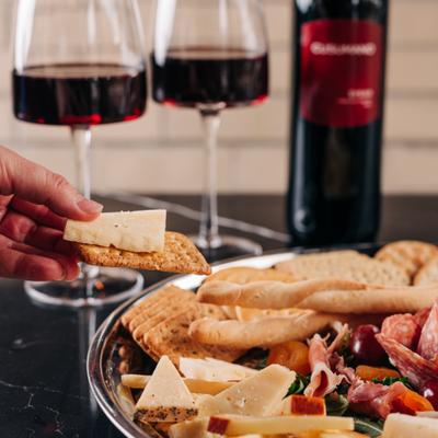 A hand taking a cracker and cheese from a charcuterie board with red wine behind.