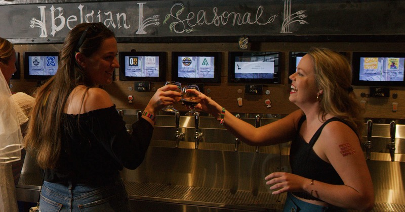 Interior, two smiling customers clink glasses in front of a beer tap wall