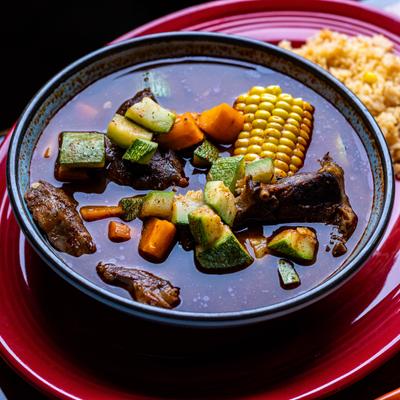 A bowl of stew with meat, zucchini, carrots, and corn on a red plate accompanied by yellow rice