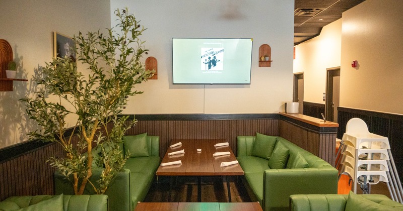 Dining area with green leather sofas and wooden tables, a large plant and a mounted TV