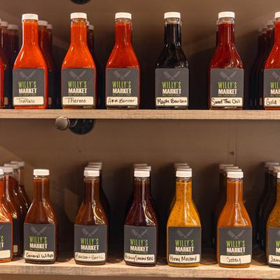 Colorful bottles of homemade sauces with Willy's Market labels arranged on a shelf.