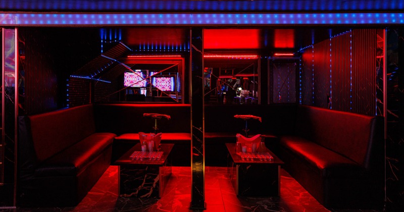A dimly lit lounge - neon lights, black leather seating, tables with ice buckets and glasses.