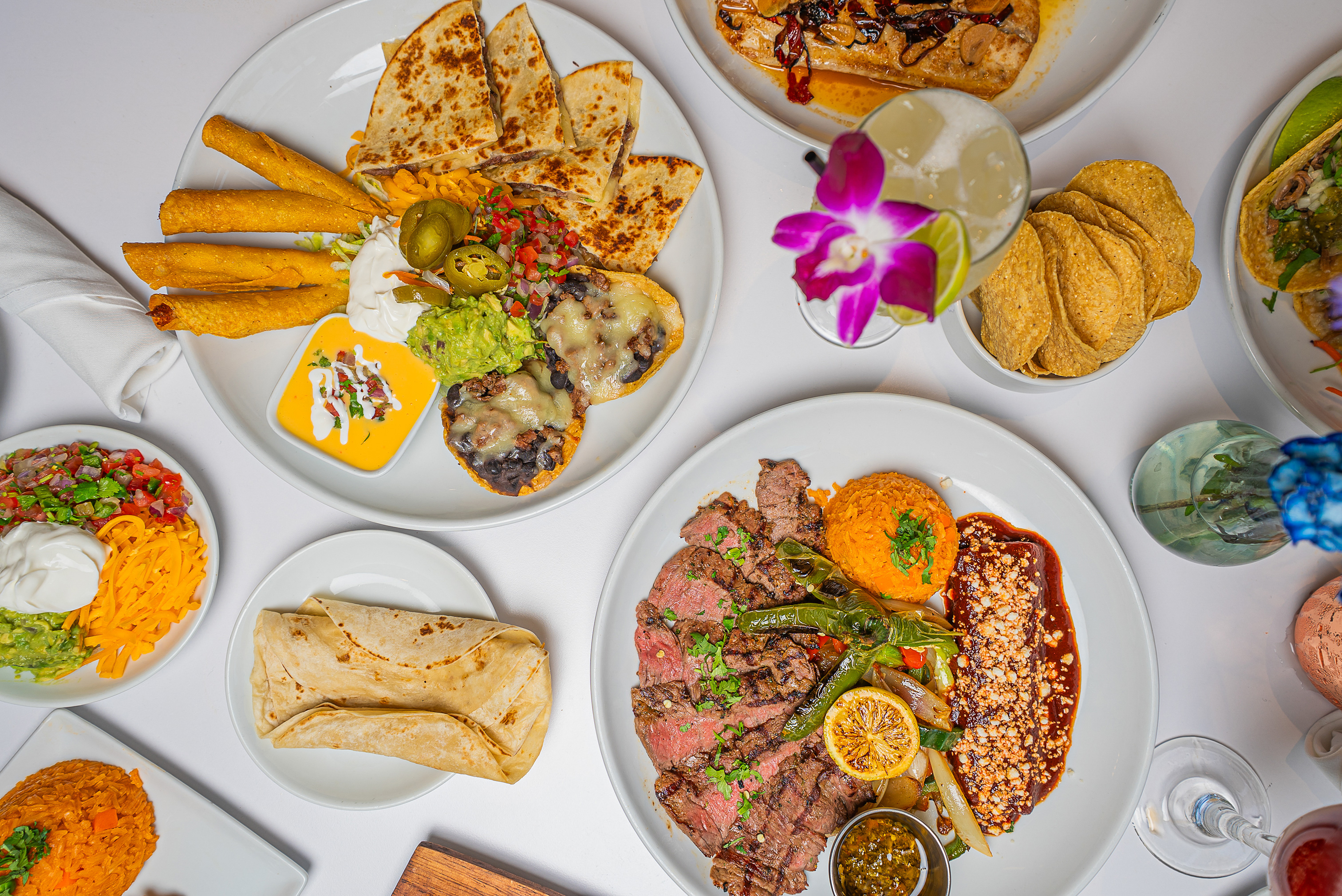 Overhead view of a dining table with tacos, steak, chips, sauces, and a cocktail