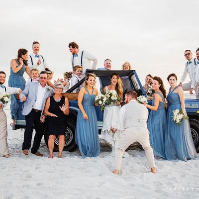 A joyful wedding party poses on a sandy beach around a blue pickup truck.