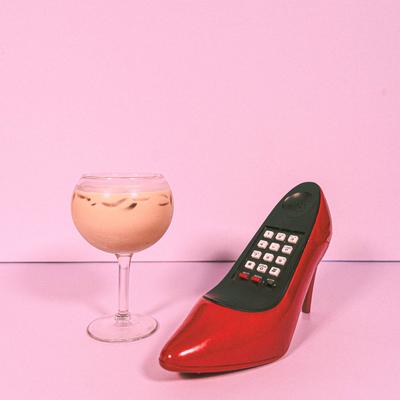 Iced cocktail in a stemmed glass beside a retro red high heel shoe telephone.