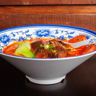 Spicy beef noodle soup with bok choy, tomatoes, scallions, and tender beef in a blue floral bowl.