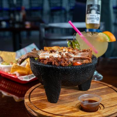 Molcajete dish served with a cocktail and a tray of chips in the background.