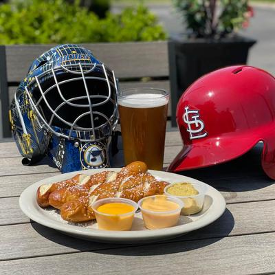 Outdoor table with served pretzel and dips, beer and a hockey and batting helmet.