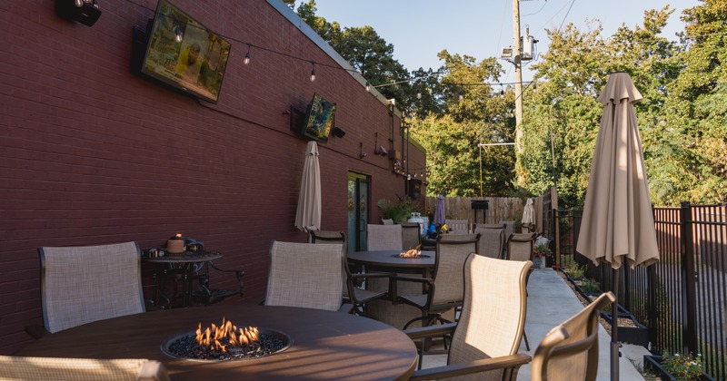 Outdoor seating area with round tables, fire pits, chairs and umbrellas next to a brick wall