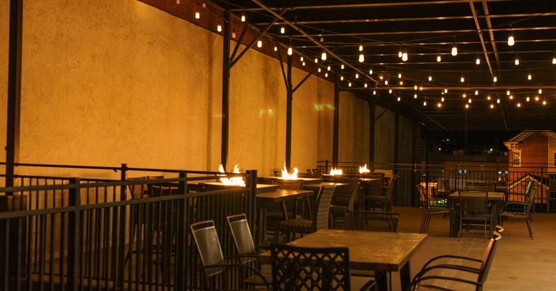 Outdoor patio with tables, chairs, string lights, and tabletop flames at night