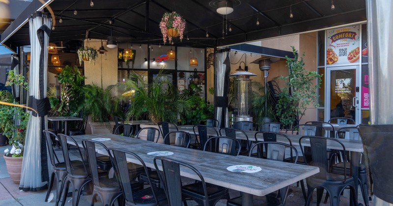 Outside, covered patio, black tables and chairs, abundant plants, hanging lights, patio heaters