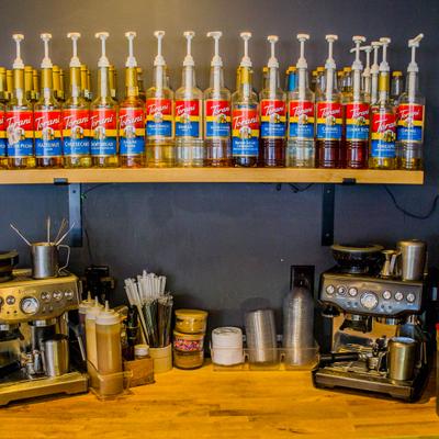 Coffee station setup featuring a wooden shelf lined with a row of flavored syrup bottles.