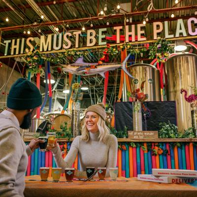 Two friends toasting with drinks in a vibrant cafe with This Must Be The Place sign.