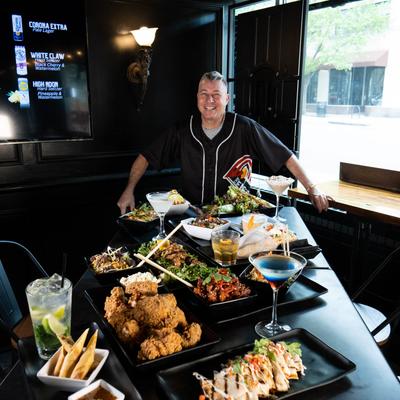 A person behind a table with assorted food and drinks, smiling at the camera.