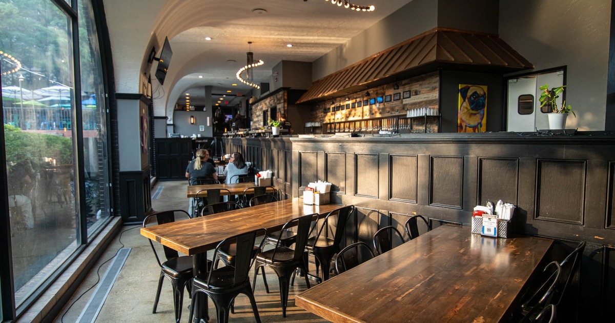 Interior with wooden tables, black chairs, and a bar, Large windows reveal greenery outside