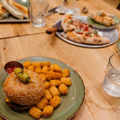 Burger with tater tots alongside pizza on a wooden table.