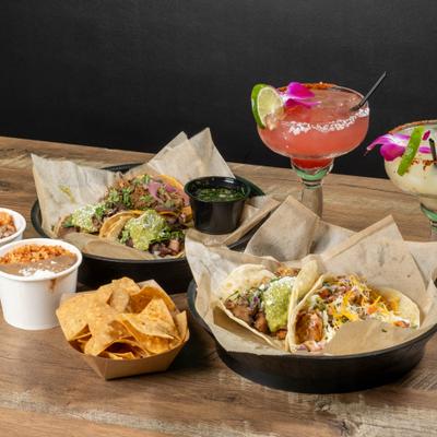 A meal consisting of tacos, side dishes, and two margaritas on a wooden table.