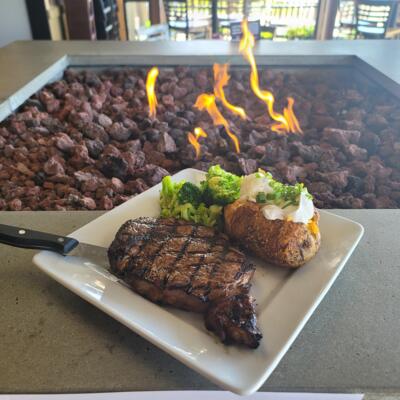 A plate with frilled steak, baked potato, and broccoli placed on the edge of a fire pit.