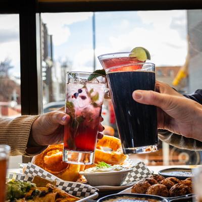 Three hands clinking glasses of colorful cocktails over a table filled with various dishes.