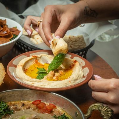A person dips pita bread into creamy hummus at a table with assorted food dishes.