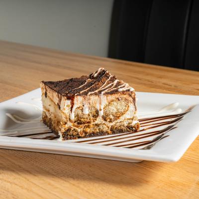 Tiramisu slice drizzled with chocolate on a white plate.