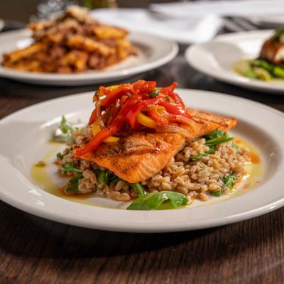 Grilled trout topped with vibrant red and yellow peppers on a bed of cooked grains and greens.