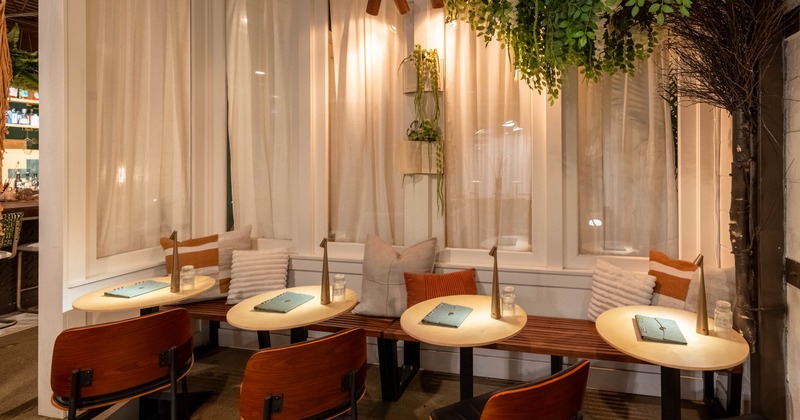 Cozy dining area with small wooden tables, wooden chairs, cushioned bench, and greenery