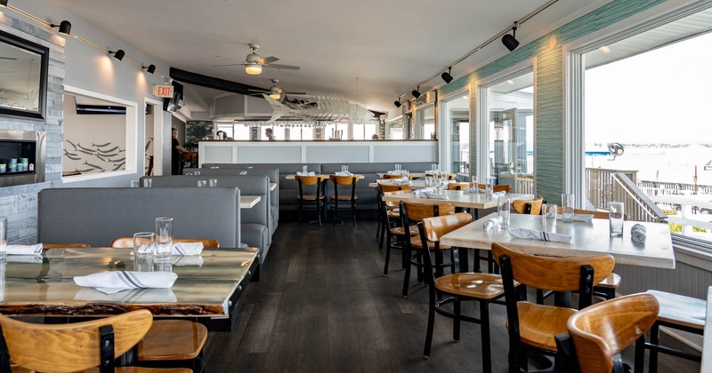 Interior of a restaurant with tables, chairs, booth seating, large windows, and nautical decor