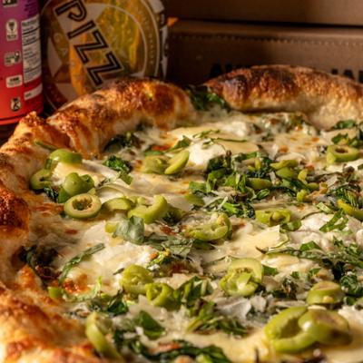 A white pizza topped with green olives, basil, and cheese next to kombucha.
