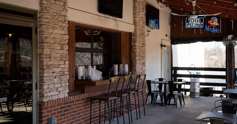 Outdoor seating area with a window counter, stools, metal tables, and beer neon signs