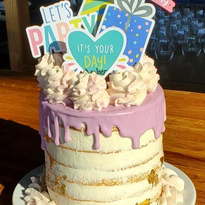 A decorated cake with pink frosting drips, topped with whipped cream and festive paper signs.