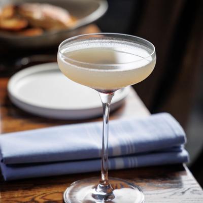 Pale yellow cocktail in a coupe glass on a wooden table beside a folded blue napkin..
