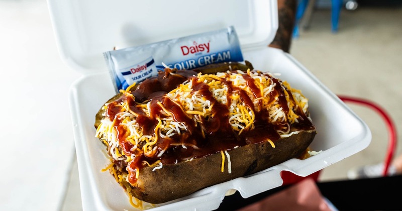 Loaded baked potato  with shredded cheese and barbecue sauce in a white takeout container
