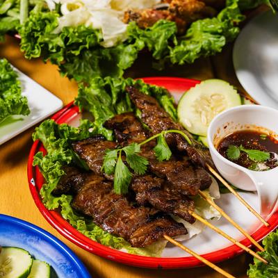 Grilled pork skewers on lettuce with dipping sauce and cucumber slices.