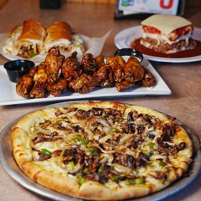 Philly Steak Pizza with chicken wings, lasagna, and sub sandwiches in the background.