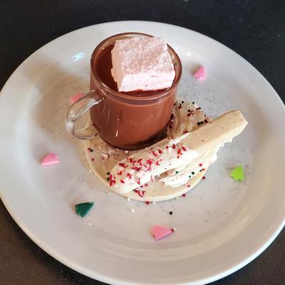 Hot chocolate glass, garnished with pink marshmallow, on a decorated plate.