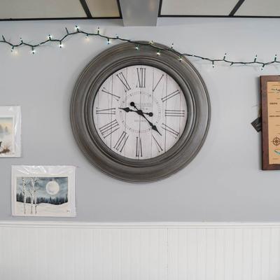 A clock with Roman numbers on a gray wall, adorned with string lights and wall art.