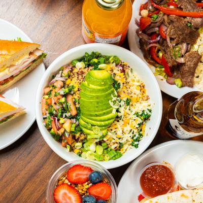 Avocado grain bowl, sandwiches, fruit parfait, grilled beef plate, and drinks on a table.