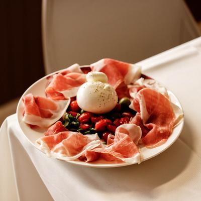 A plate of burrata with prosciutto, tomatoes, olives and basil on a white tablecloth.