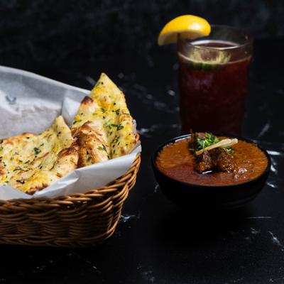 Naan basket, a bowl of curry dish, and a glass of raspberry lemonade on a dark table.