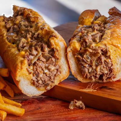 Two cheesy steak sandwiches on a wooden board with fries.