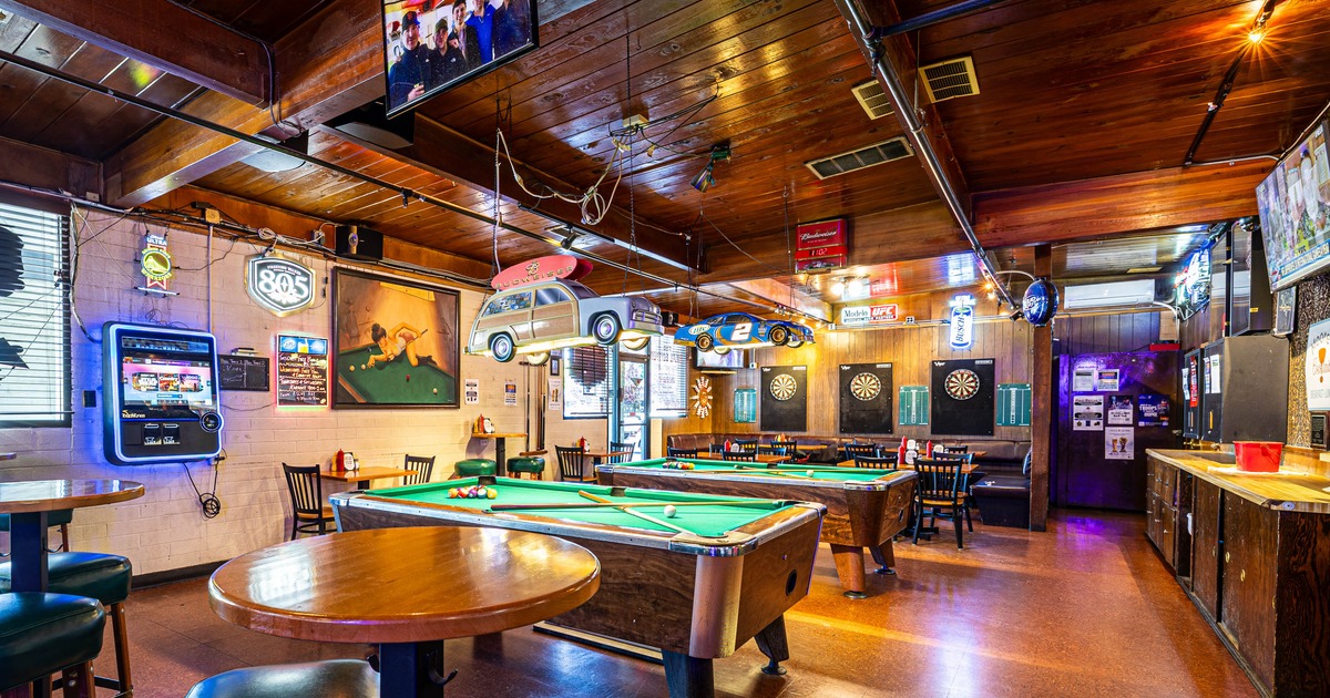 A rustic bar interior - a pool table, dartboards, featuring wooden decor, neon signs and model cars