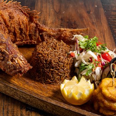 Whole fried fish with black rice, salad, plantains, and lemon garnish.