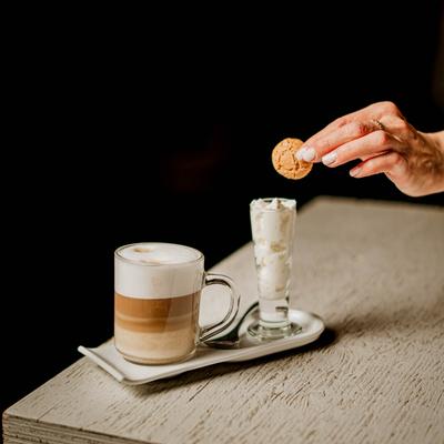A person dips a cookie into a glass of whipped cream served next to a glass of latte.