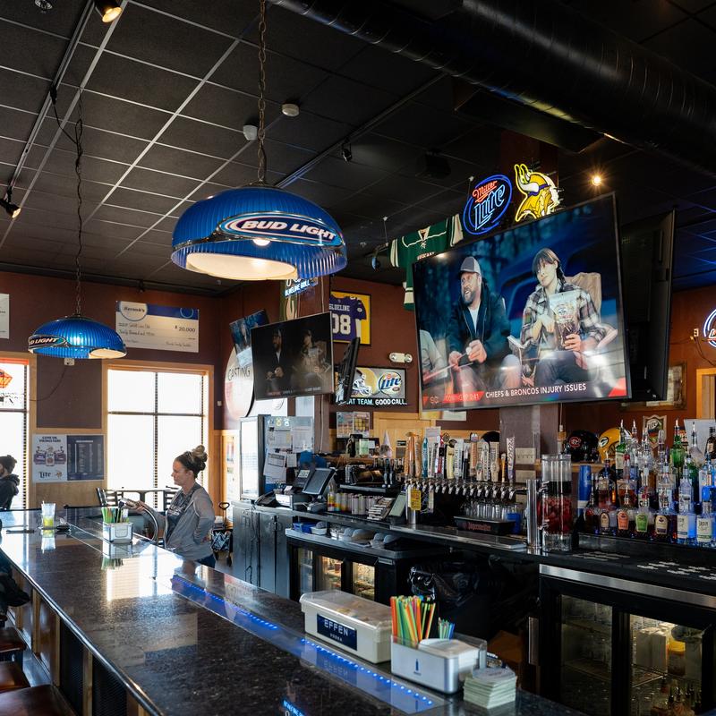 Interior of a bar with a bartender, patrons, liquor bottles, and TVs displaying sports broadcasts