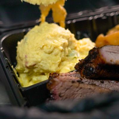 Smoked BBQ brisket with creamy potato salad in a black takeout container.
