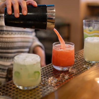Cocktial being poured from the shaker into a glass, alongside other cocktails.